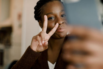 Teenage girl showing peace sign while taking selfie through smart phone at home