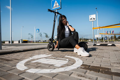 Portrait of woman with push scooter sitting on footpath against blue sky