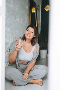 Beautiful young woman plus size body positive in comfortable sport wear with glass of water at home