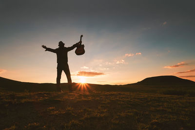 Silhouette man with guitar standing on land against sky during sunset