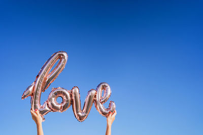 Crop anonymous woman holding silver inflatable balloon in shape of word love against cloudless blue sky in sunny summer day