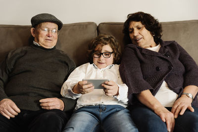 Grandparents looking at grandson using smart phone while sitting on sofa at home