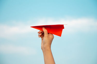 Cropped hand holding red paper plane against sky