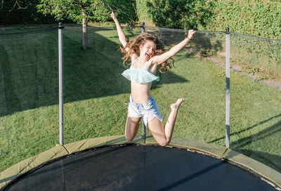 Horizontal photo of a smiling young brunette teenager jumping on a trampoline with net around on the green yard outdoors. the girl wears short jeans and a top and looks fun, active