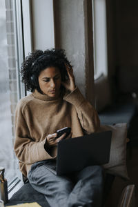 Female entrepreneur wearing wireless headphones and working on laptop by window in creative office