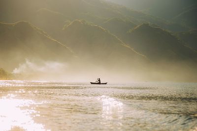 Silhouette man rowing boat on river against mountains