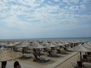 Thatched parasols and deck chairs at beach against cloudy sky