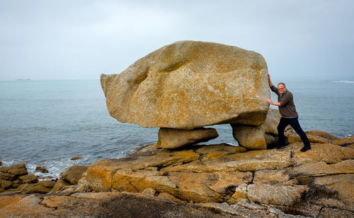 Man standing pushing rock by sea against sky