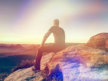 Tourist man sit on stone at cliff edge overlooking misty land to horizon. rear view. abstract light