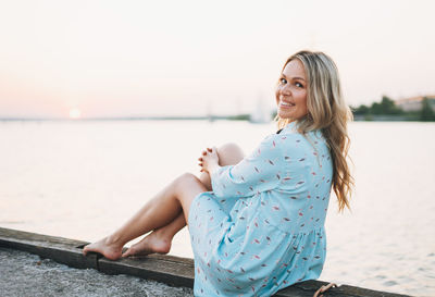 Beautiful blonde young woman in blue dress sitting on pier and looking on sunset