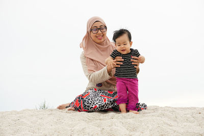 Mother with daughter sitting on sand at beach against clear sky