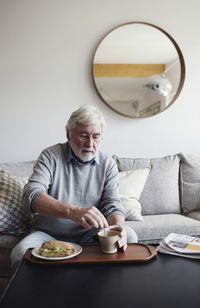 Senior man stirring tea while having breakfast at table in living room