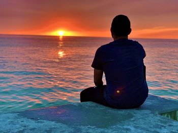 Rear view of man sitting on sea against sky during sunset