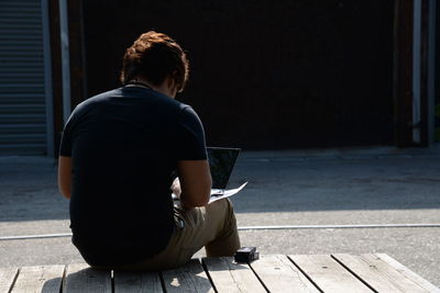 Rear view of man using laptop while sitting on wooden bench