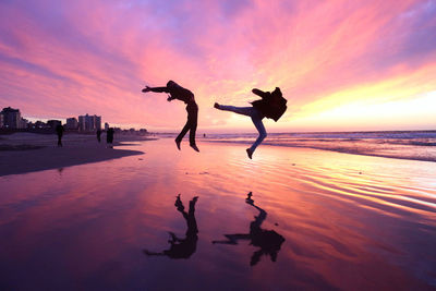 People jumping at beach against dramatic sky during sunset