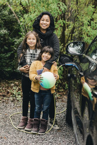 Portrait of smiling woman with daughters by electric car at charging station