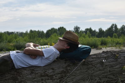 Side view of young man with hat and backpack lying on tree trunk against cloudy sky