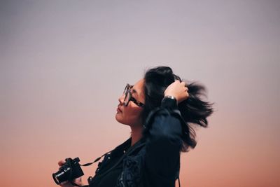 Young woman with hand in hair holding camera while standing against sky during sunset