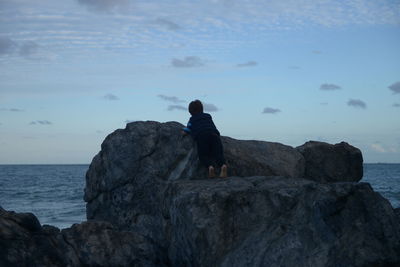 Rear view of boy standing on rock at beach against sky
