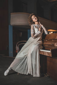 Full length of sensuous woman in white dress sitting on piano at home