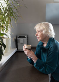 An attractive elderly woman drinks espresso in the kitchen and looks out the window.