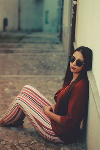 Portrait of a young  woman sitting against  wall wearing sunglasses bourdeous shirt and stripes pant