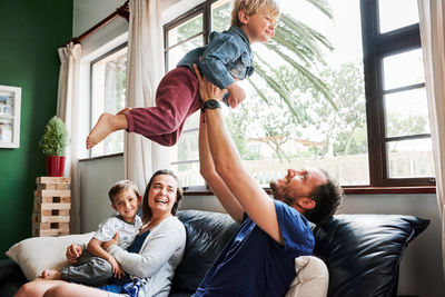 Cheerful father holding son mid air while sitting on sofa at home-