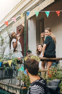 Happy young women waving hands at male friend while standing in balcony during party