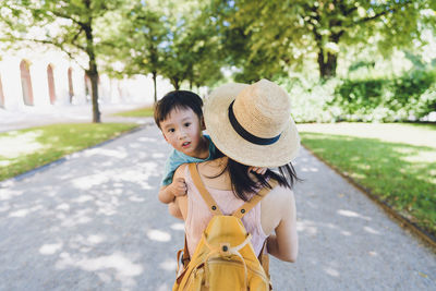 A boy looks back at the camera while his mother holding him walking toward a park.