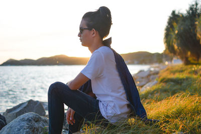 Side view of young man relaxing on field by sea against sky during sunset