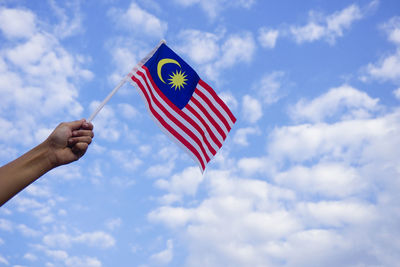 Cropped hand holding malaysian flag against cloudy sky