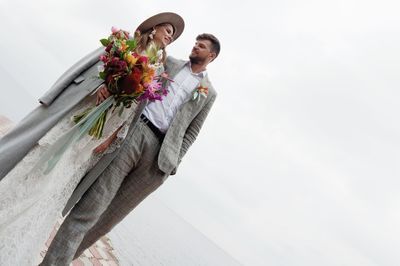 Newly married couple walking on pier over lake against cloudy sky