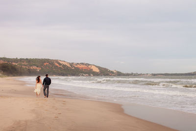 Rear view of couple walking at beach against sky