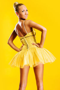 Rear view portrait of beautiful ballerina standing with hands on hip against yellow background