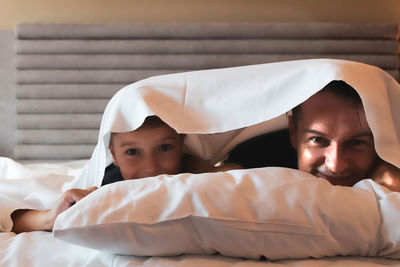 Happy boy and his father relaxing on abed while being covered with bedsheet over their heads.