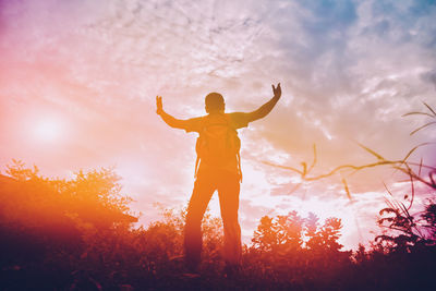 Rear view of man with arms raised standing against sky during sunset