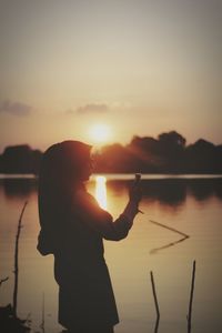 Woman photographing lake while standing against sky during sunset