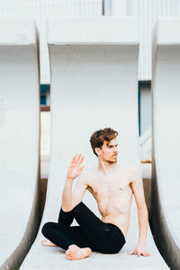 Shirtless man practicing yoga while sitting on built structure