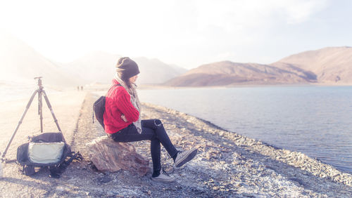 Side view of woman sitting at lakeshore against mountains