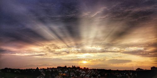 Sunlight streaming through silhouette buildings against sky during sunset