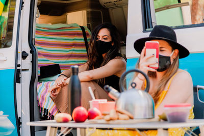 Calm female friends wearing protective masks sitting near van and enjoying picnic during coronavirus epidemic