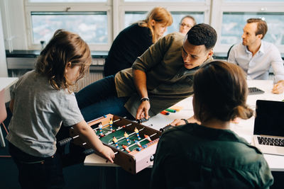 High angle view of boy playing foosball with businessman on desk amidst colleagues at creative office