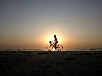 Silhouette woman riding bicycle at beach against sky during sunset