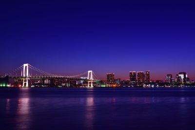Illuminated bridge over river by buildings against clear blue sky at night