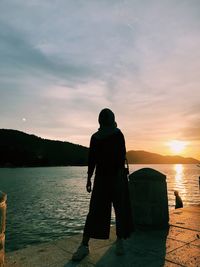 Rear view of man standing by lake against sky during sunset