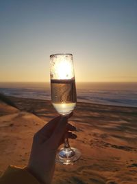 Hand holding glass of water at beach against sky during sunset