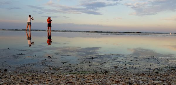 Full length of woman photographing man and baby at beach against sky during sunset