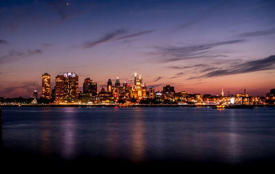 Illuminated buildings by sea against sky at dusk