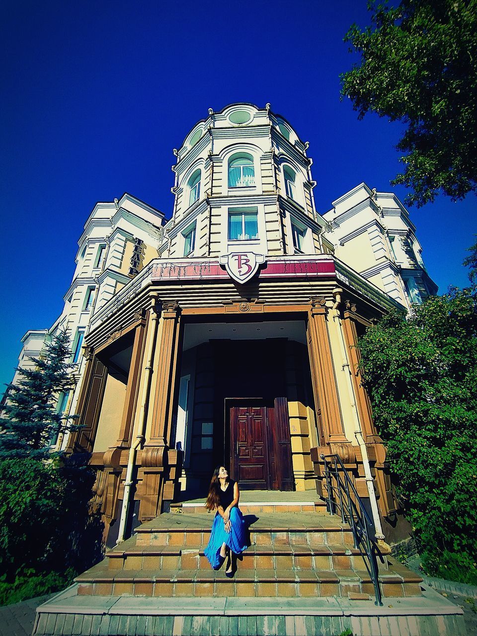 REAR VIEW OF WOMAN SITTING AGAINST BUILDING AGAINST BLUE SKY