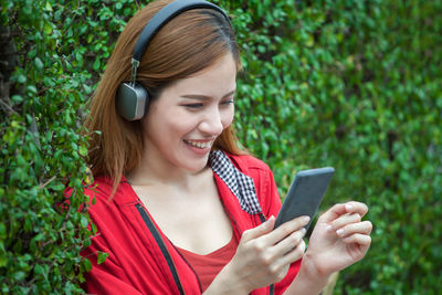 Smiling beautiful woman listening to music while leaning on hedge in garden
